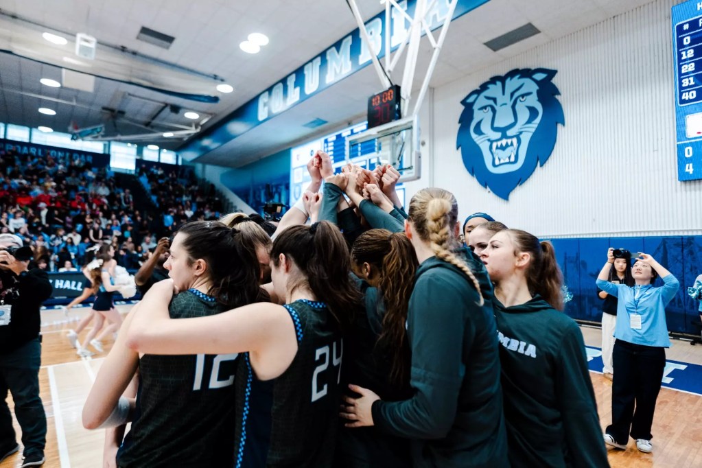 Columbia players huddle during a game. Some of them are wearing dark gray jerseys with light blue trim, and others have dark gray warmup shirts on. A clock in the background shows 10 minutes.