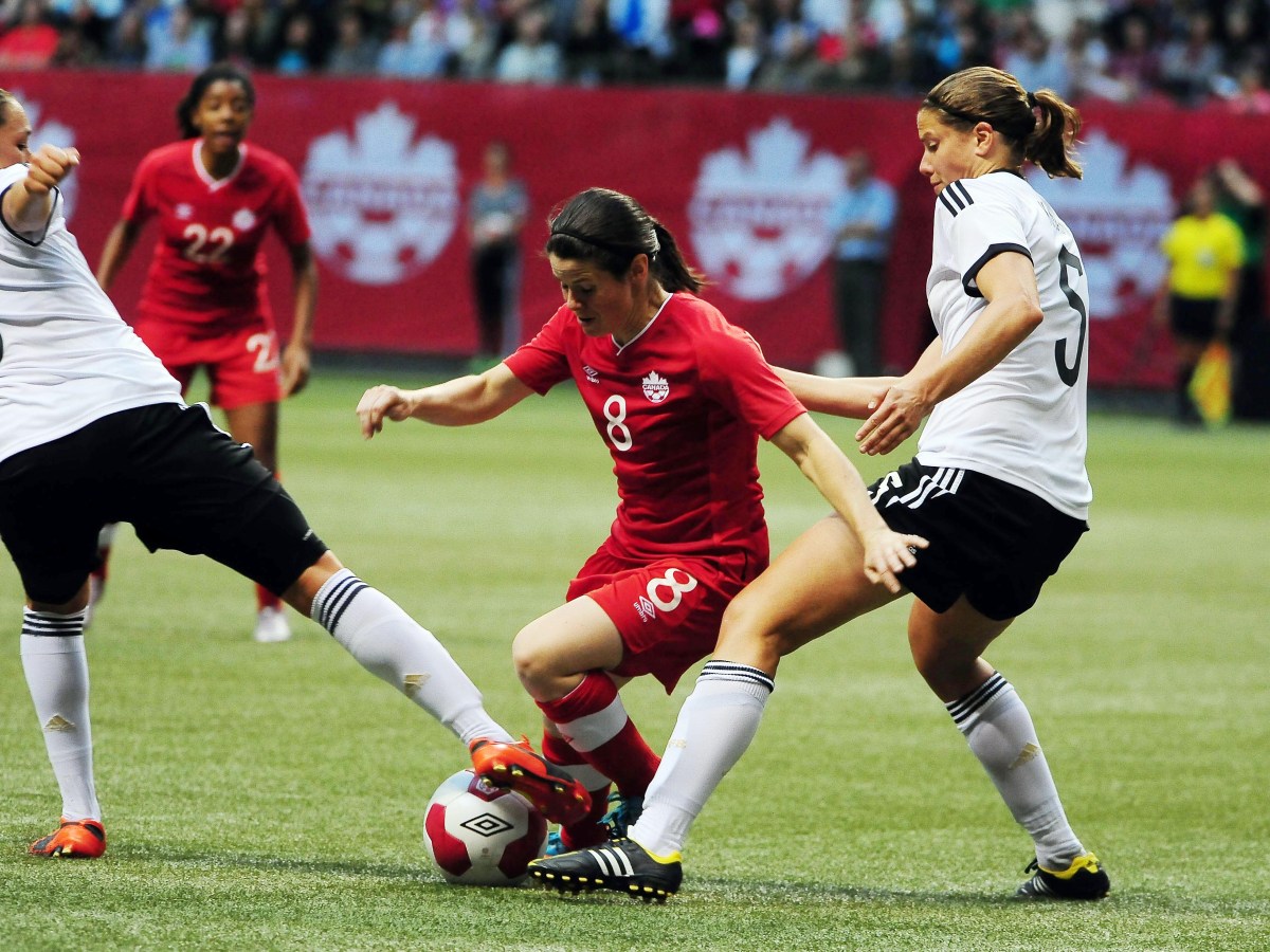 Diana Matheson battles for the ball against Nadine Kessler and Annike Krahn. The Canada Soccer logo appears multiple times on boards beyond the field.
