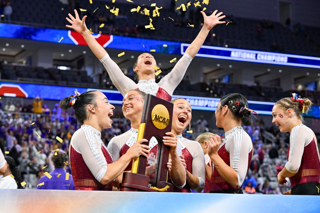 A group of gymnasts in University of Oklahoma leotards hold the national championship trophy together and yell in celebration.