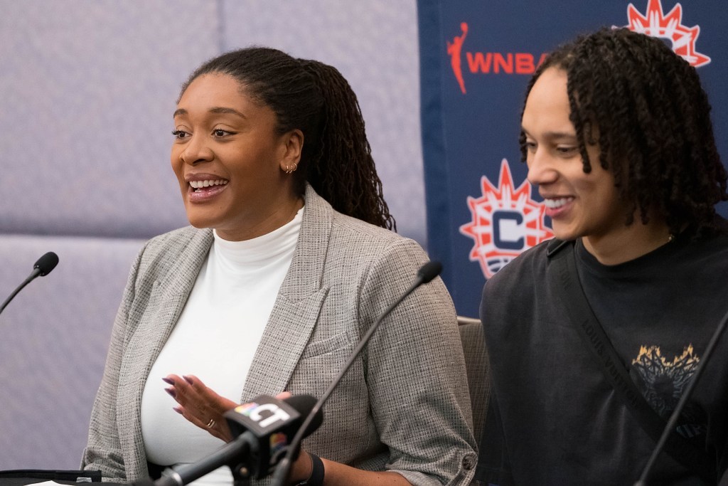 Morgan Tuck and Brittney Griner sit next to each other at a press conference. Tuck wears a blazer and a white shirt and Griner wears a black sweatshirt.