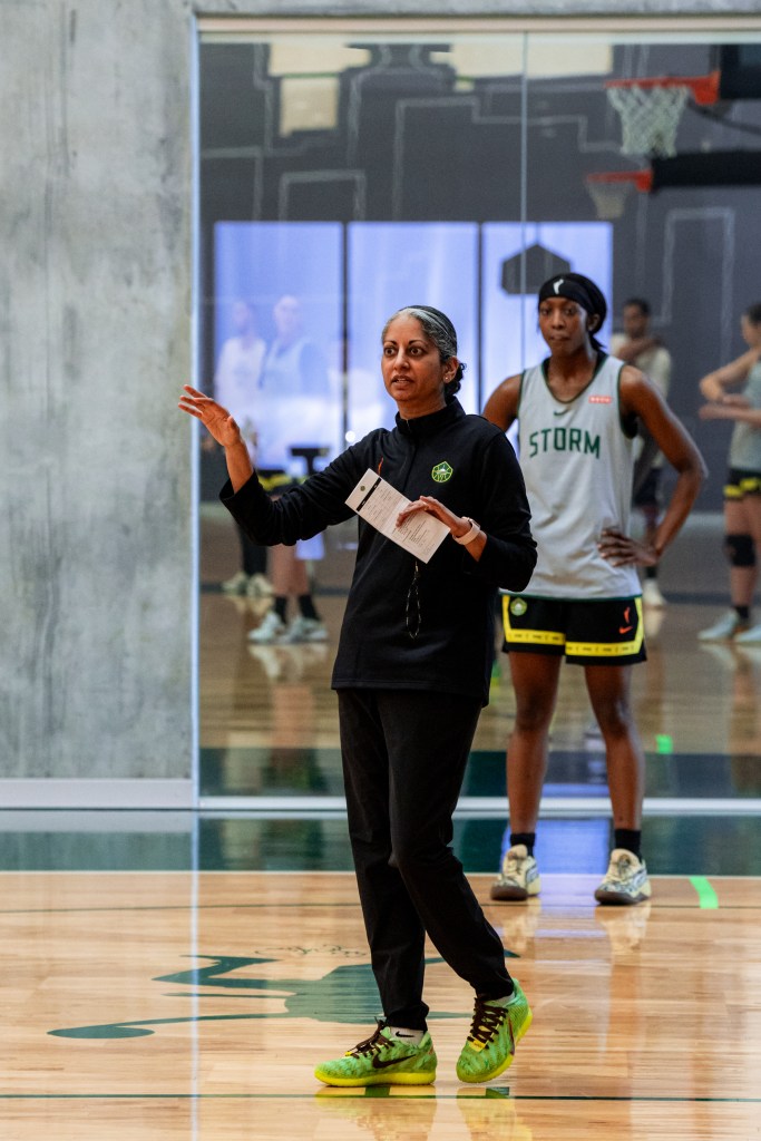Head coach Sonia Raman holds a piece of paper and stands on the court explaining something with her other hand while Flau'jae Johnson looks on from several feet behind with a focused expression.