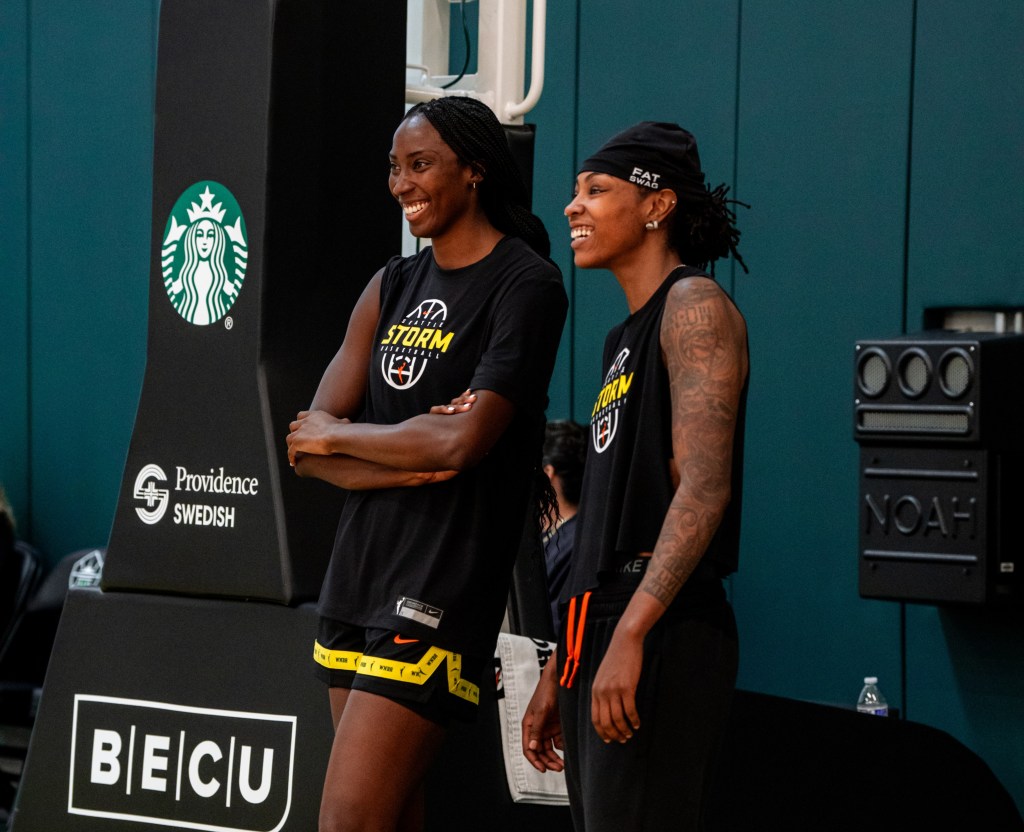 Ezi Magbegor stands with her arms crossed and a wide smile on her face next to Jordan Horston whose arms are relaxed but her face is similarly smiling.