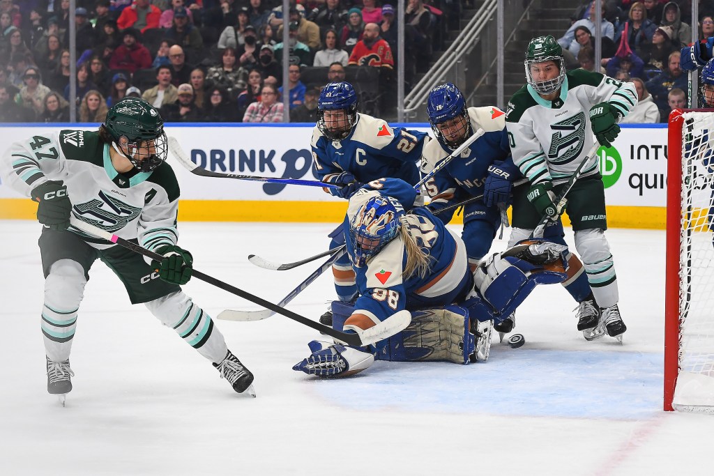 Rattray watches the puck go in after taking a backhand shot, while players battle in the background. She is wearing a white away uniform.