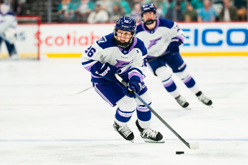 Coyne Schofield looks up ice as she skates with the puck. She is wearing a white away uniform.