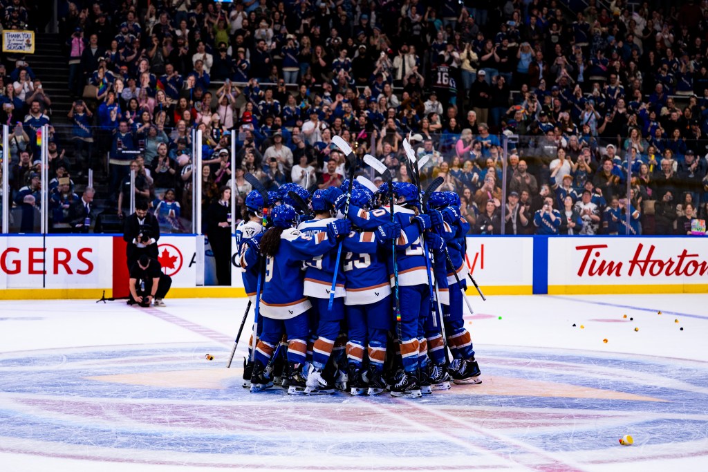 The Goldeneyes celebrate with a tight group hug at center ice, as rubber ducks litter the ice, thrown by fans in celebration. They are wearing blue home uniforms.