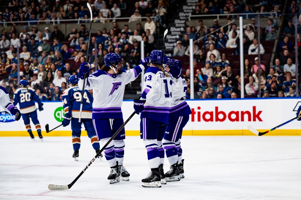 Frost players celebrate a goal against the Goldeneyes with a group hug. They are wearing white away uniforms.
