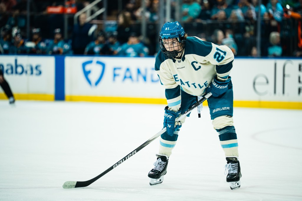 Knight leans on her stick and looks into the circle, waiting for the puck to drop. She is wearing a cream away uniform.