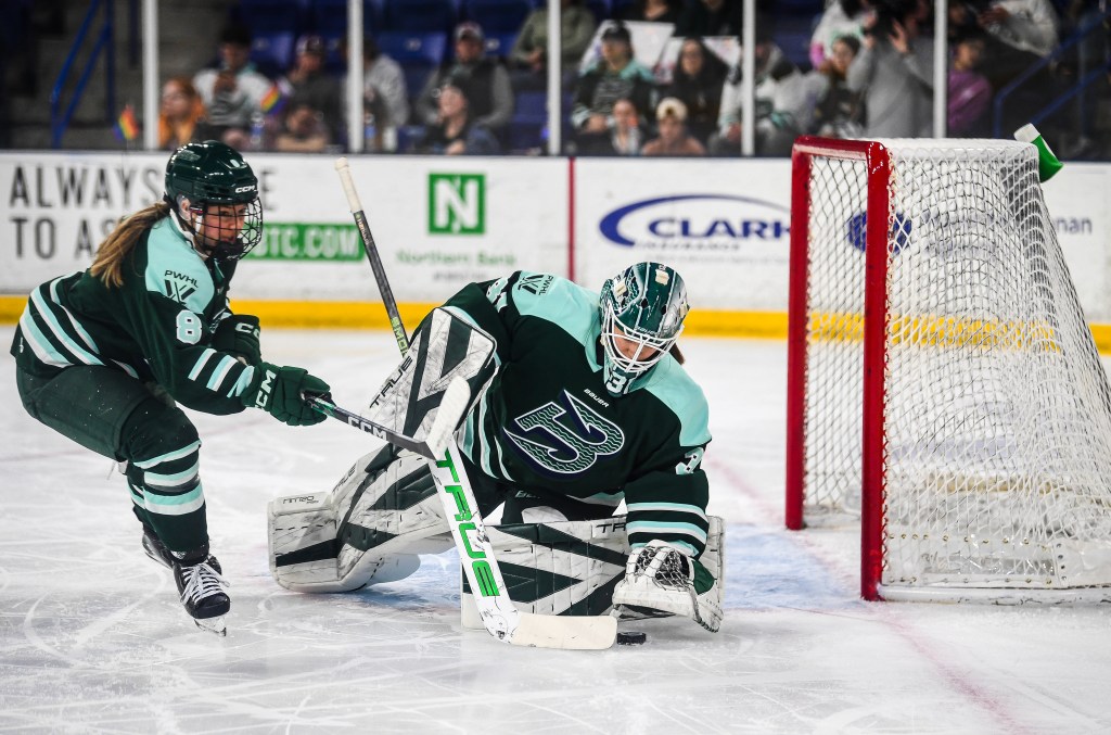 Frankel covers the puck with her glove after making a save on he rknee, while Winn backs her up. They are wearing green home uniforms.