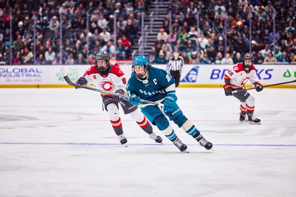 Knight skates after the puck with a Charge playing trailing her. She is wearing a teal home uniform, while the Charge player is in a white away one.