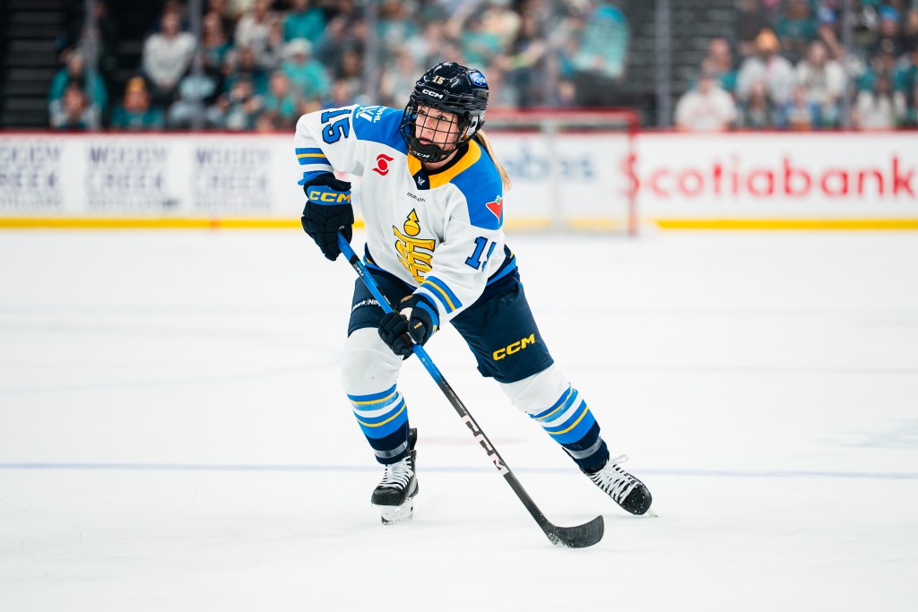 Harmon looks up ice as she skates with the puck. She is wearing a white away uniform.