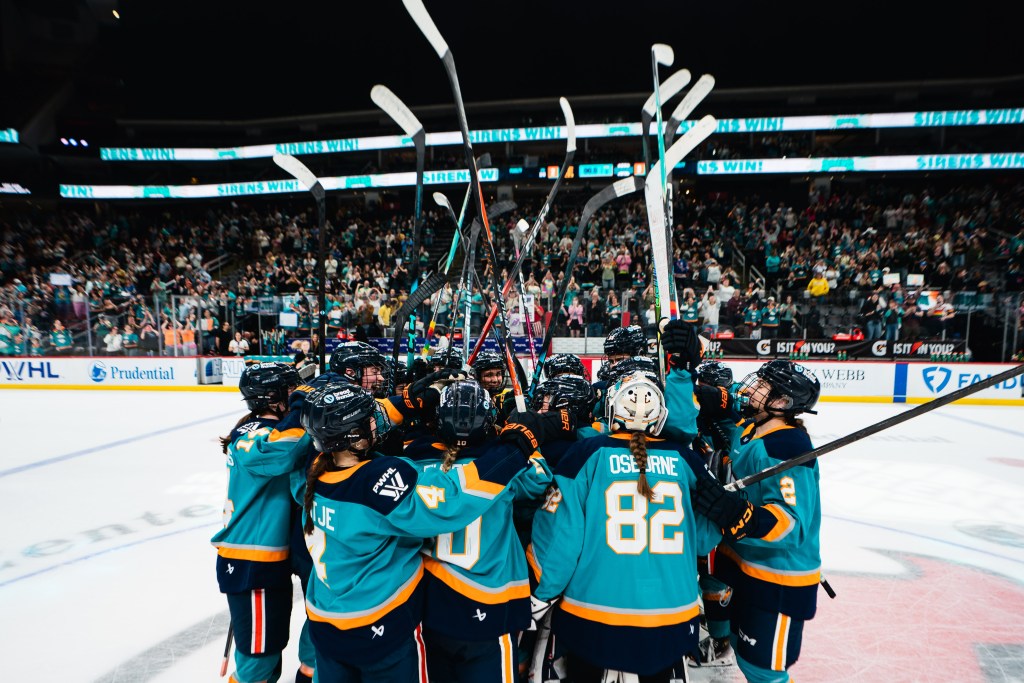 The Sirens raise their sticks and come together for a tight group hug at center ice. They are all wearing teal home uniforms.