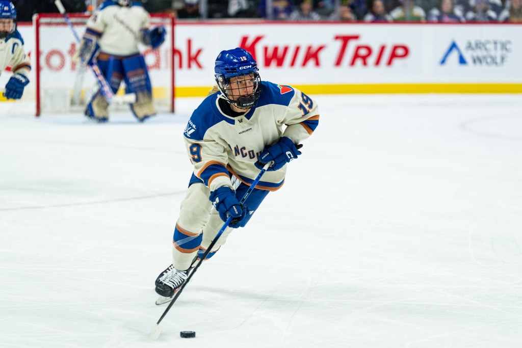 McMahon looks up ice as she skates the puck out of Vancouver's defensive zone. She is wearing a cream away uniform.