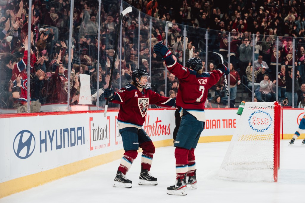 With arms open wide, Scamurra and Stacey come together for a hug. They are wearing maroon home uniforms.