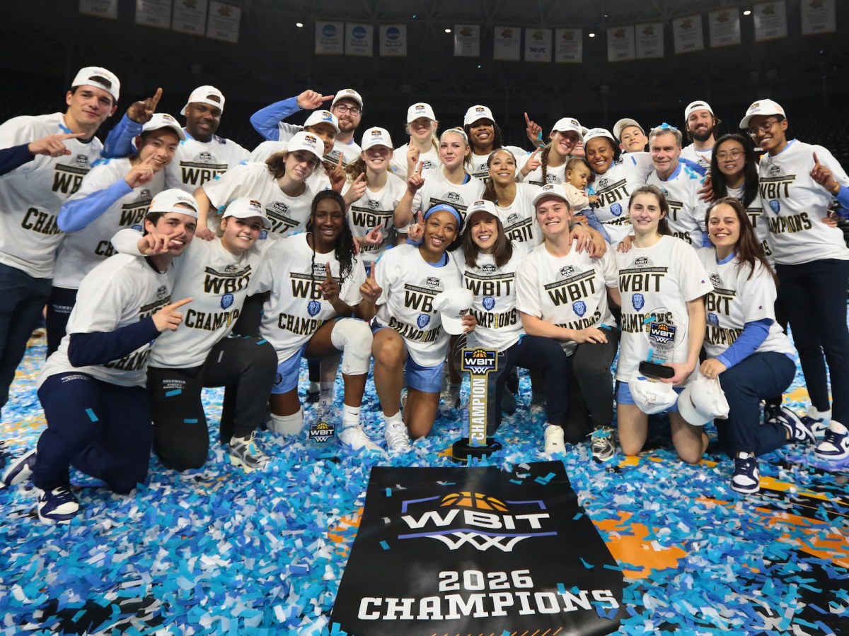 Columbia players and coaches smile for a photo on a basketball court. A generous amount of light blue and white confetti is coating the ground, and everyone is wearing white T-shirts that say "WBIT champions." The WBIT trophy and a commemorative towel are displayed in front of them.