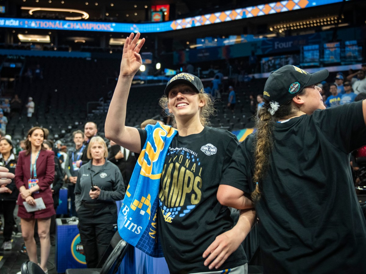 UCLA guard Gianna Kneepkens smiles and waves as she celebrates the Bruins' NCAA championship. She is wearing a black championship hat and a black shirt that reads, "National champs." She also has a blue UCLA towel draped over her right shoulder.