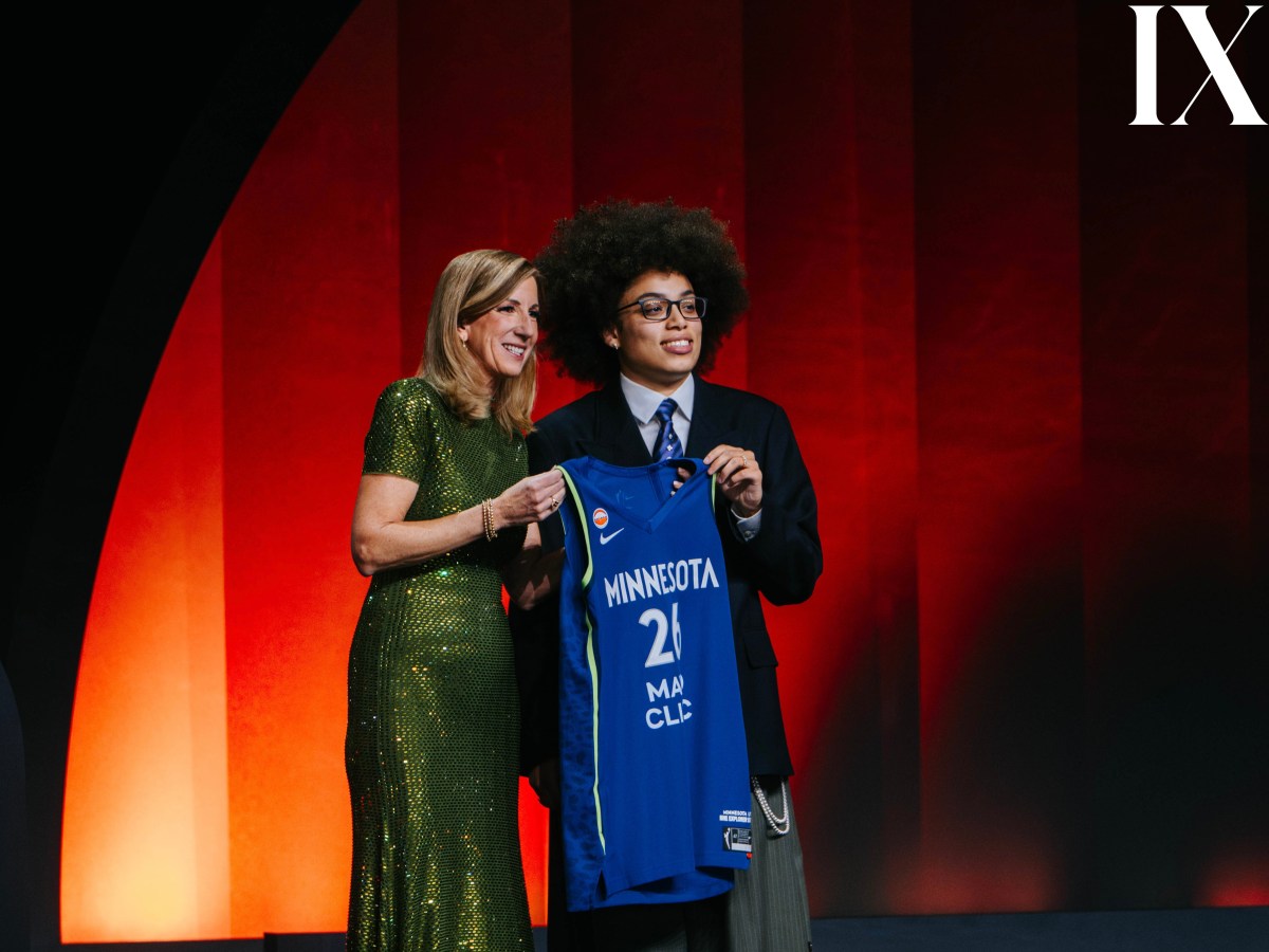 Olivia Miles (right) holds up a blue Minnesota Lynx jersey on a bright stage with commissioner Cathy Engelbert wearing a green dress. Miles is wearing a dark jacket with a big smile on her face.