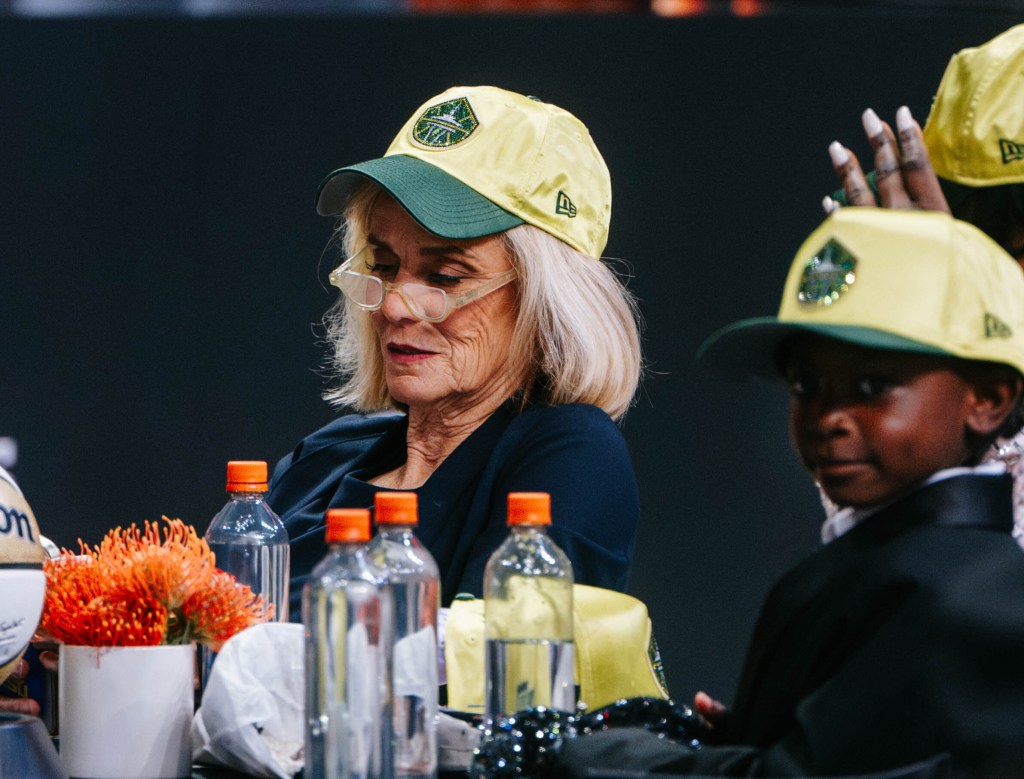 Kim Mulkey looks down at her phone through her glasses with a concentrated look. Flau'jae Johnson's little brother looks directly at the camera shooting from a distance. Both sport Seattle Storm hats.