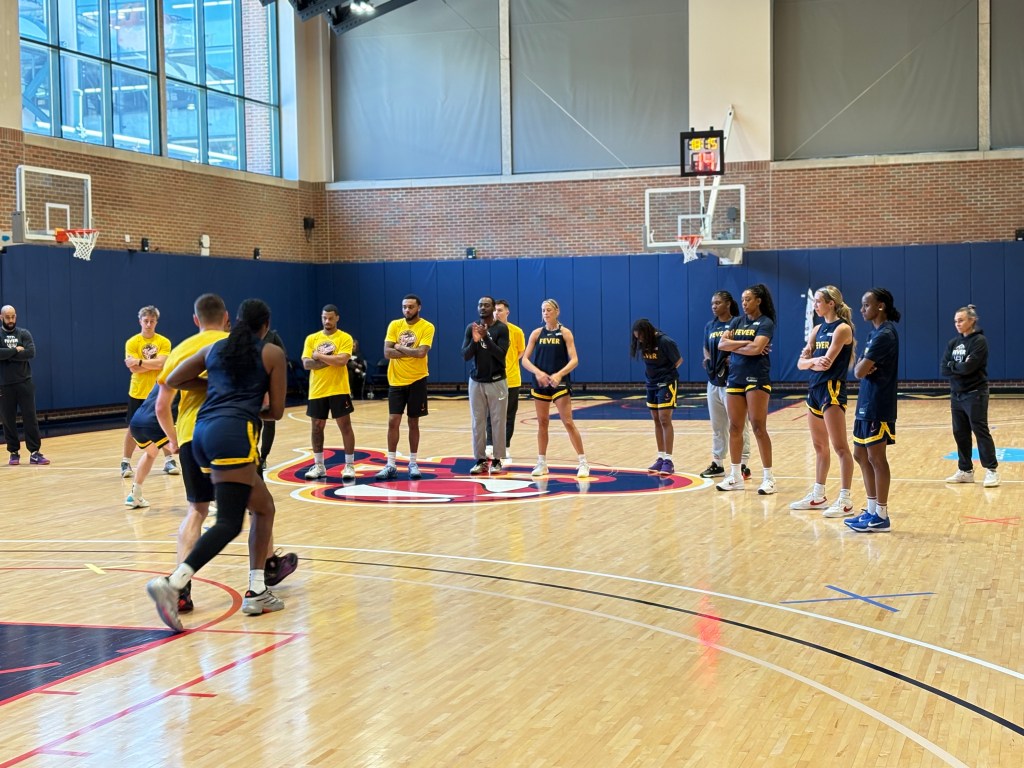 Indiana Fever players stand at halfcourt on a practice court, watching the team in defensive drills and defending practice players. The players are in navy blue Fever tshirts while all the practice players are scattered about in yellow Fever tshirts