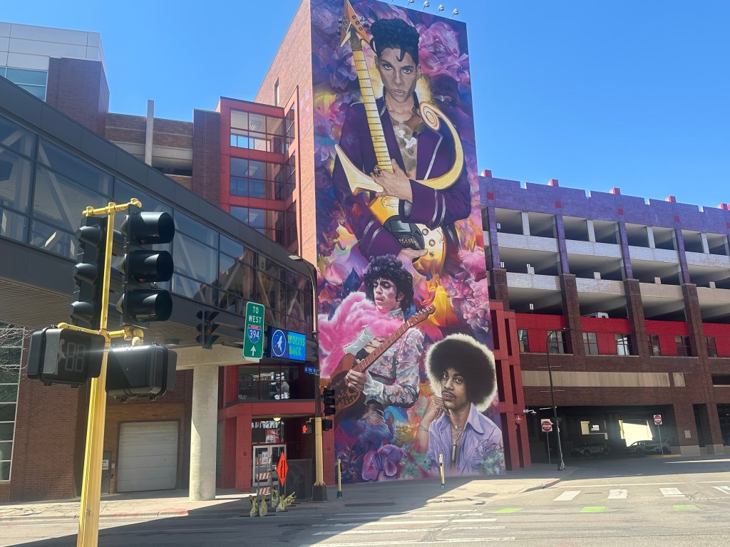 A pillar on the side of a building has a purple mural of Prince at three different stages of his life painted on it.