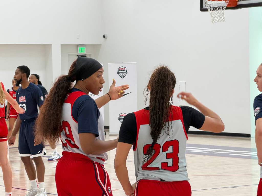 Basketball players in white and red uniforms discuss strategy on court. One gestures while speaking. Intense focus, indoor gym setting, USA logo visible.