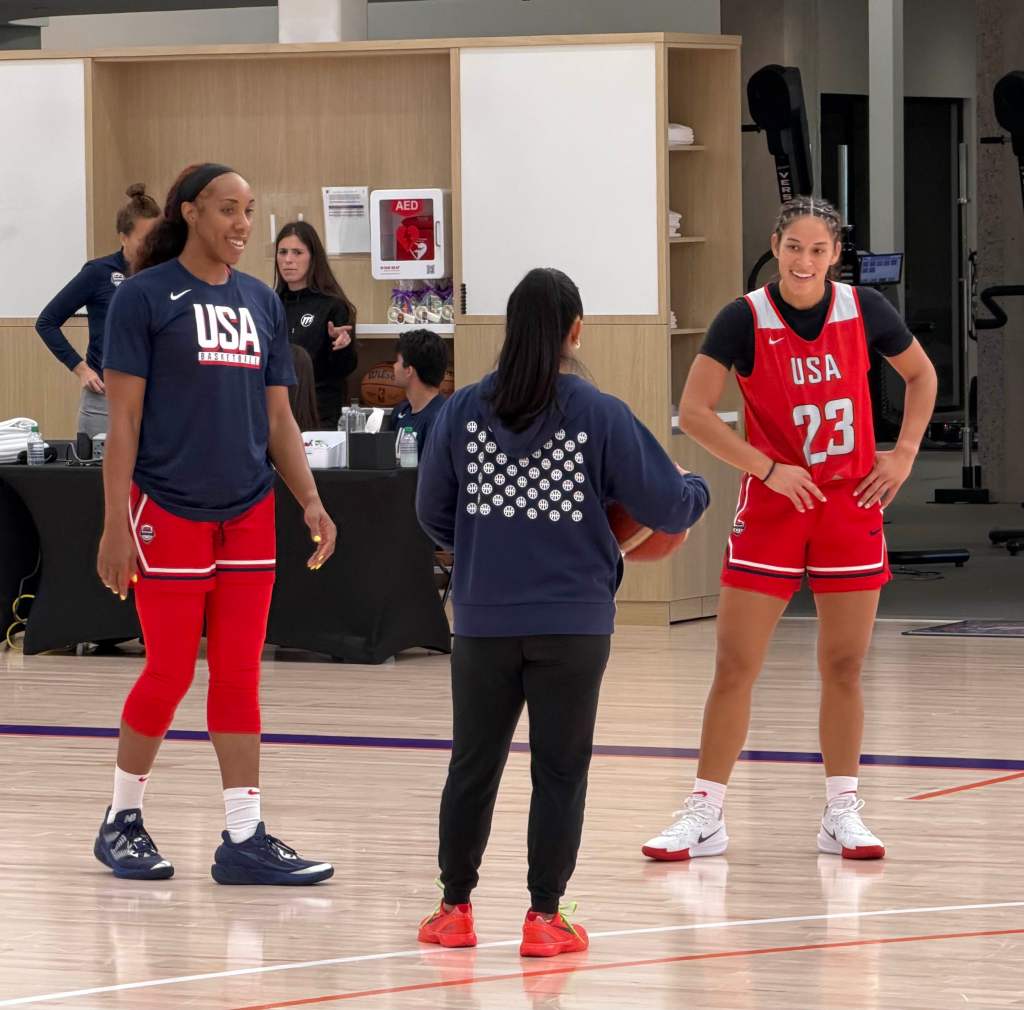 Two female basketball players wearing USA uniforms smile during practice while listening to a coach holding a ball. Energetic and focused atmosphere.