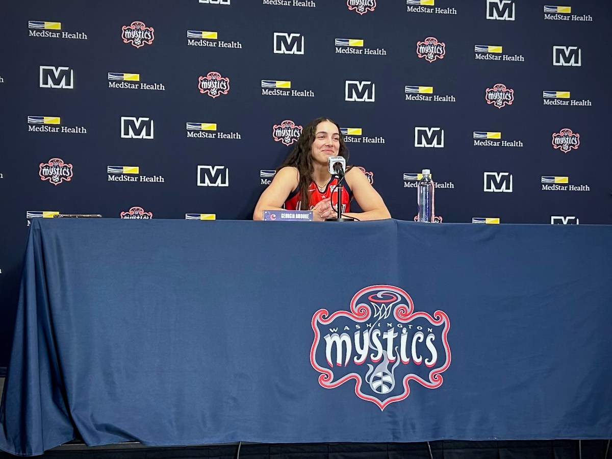 Washington Mystics point guard Georgia Amoore sits at a table and smiles as she listens to a question from the media. She's wearing a red Mystics jersey, which pops against the navy backdrop and navy tablecloth. Both the backdrop and the tablecloth have Mystics logos on them.