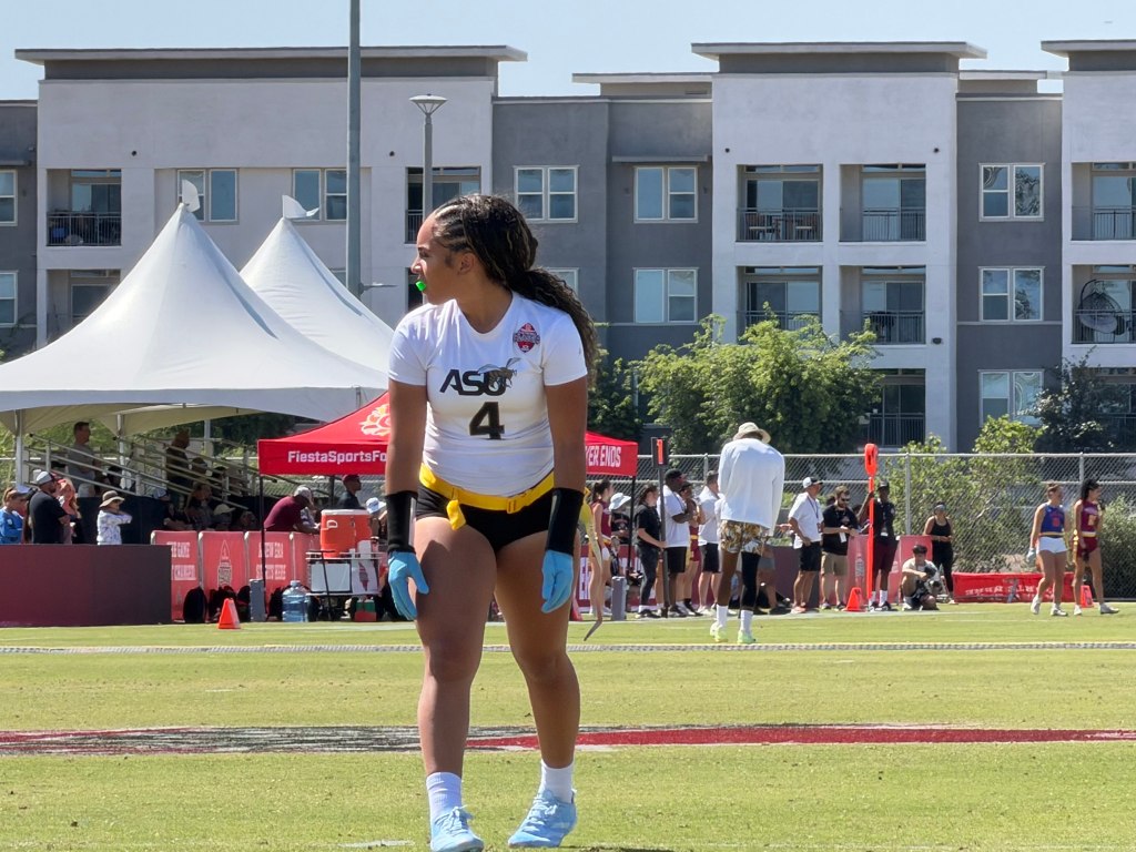 A female flag football player wearing a white jersey with the number 4 stands on a sunny field. In the background, there are white tents and spectators.
