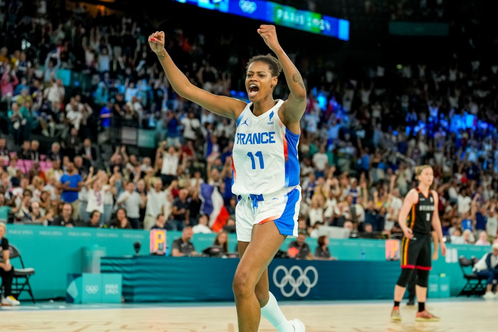 Valeriane Ayayi in a France jersey jubilantly raises arms on a court, celebrating. A cheering crowd and Olympic logo are visible in the background.