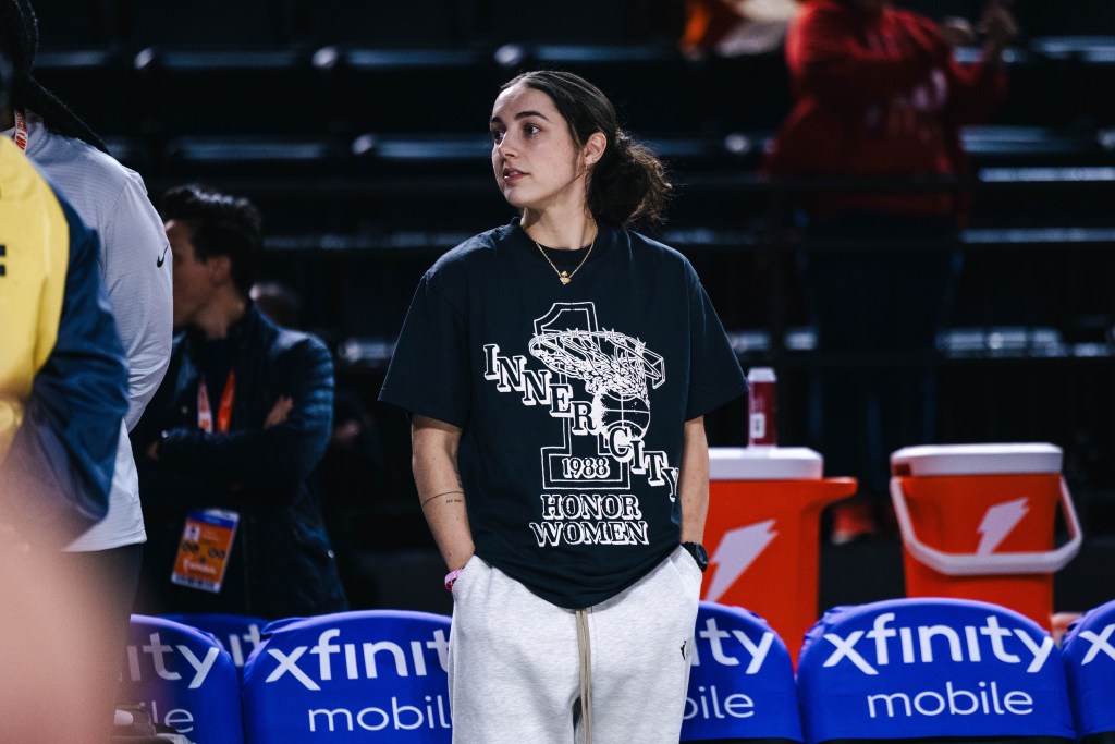 Washington Mystics point guard Georgia Amoore stands near the bench, wearing a black T-shirt and gray sweatpants. She has her hands in her pockets and is looking off to her right, watching warmups out of the frame.
