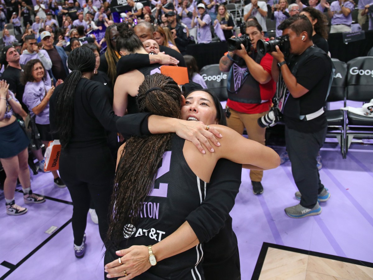 A basketball player in a black and purple jersey hugs a coach on a crowded court. Fans and photographers capture the joyful, celebratory moment.