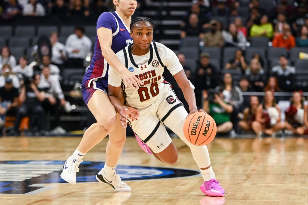Ta'Niya Latson in a South Carolina uniform drives past a defender on a court, holding the ball with focus and determination. Crowd in background.