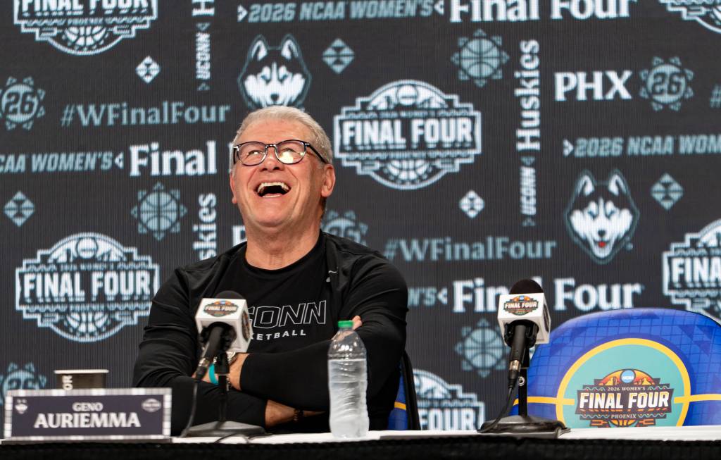 Geno Auriemma at a press conference laughs heartily, sitting at a table with microphones. The backdrop features "Final Four" logos and team graphics.