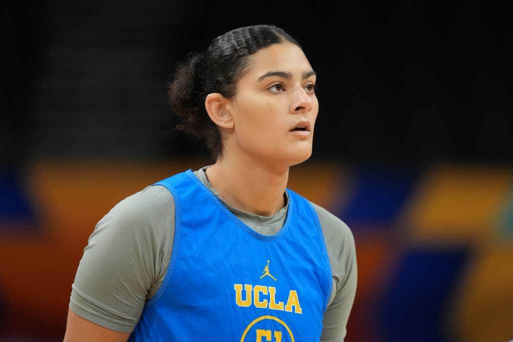 Lauren Betts, wearing a blue UCLA jersey and gray undershirt, gazes attentively. The background is a blurry arena setting.