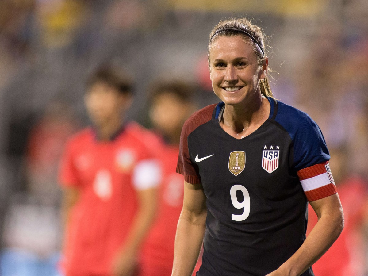 Heather O'Reilly smiles in a dark USWNT kit. She wears the captain's armband.