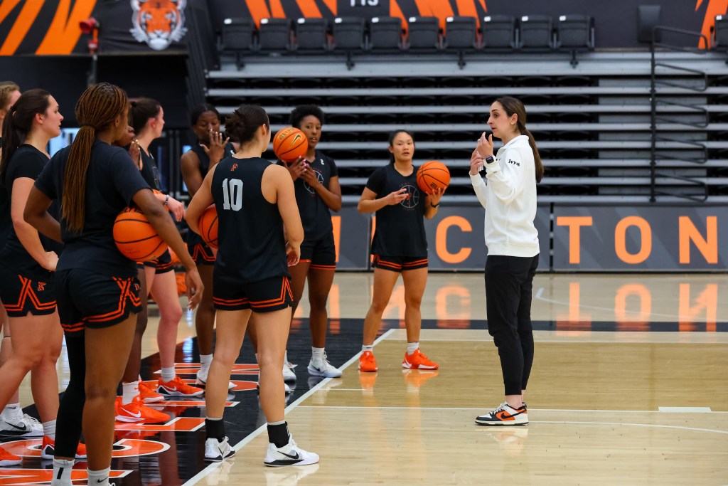 Princeton players stand in a semi-circle on the baseline during practice and listen to head coach Lauren Gosselin talk. Each player is holding a basketball.