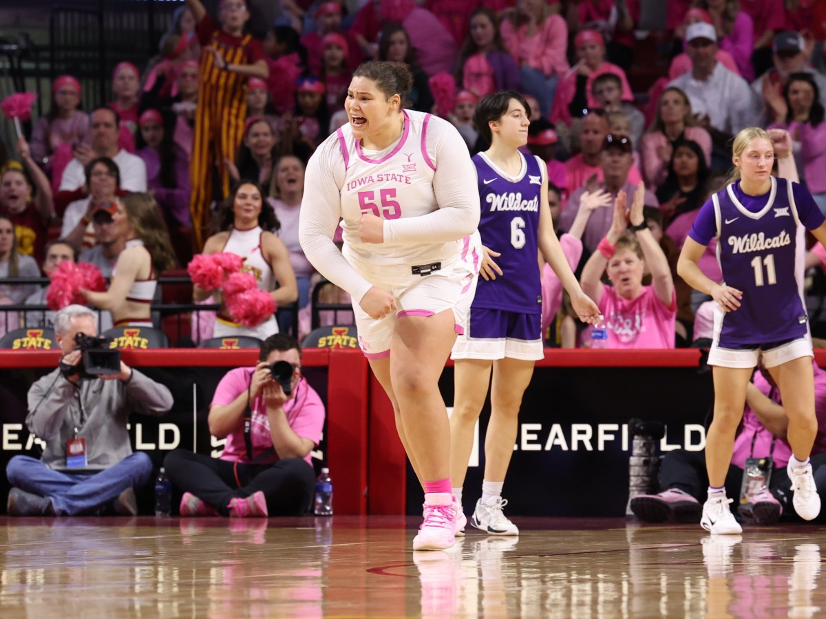 Audi Crooks flexes running back on defense. She is wearing a white Iowa State jersey with pink piping and a Play for Kay logo. Two Kansas State players are in the background in purple jerseys looking to get the ball up the court.