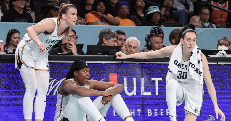 Sabrina Ionescu, Jonquel Jones and Breanna Stewart all wait at the scorers table to check into a game. Ionescu is standing, Stewart is kneeling, and Jones is seated between them with her elbows resting on her knees.