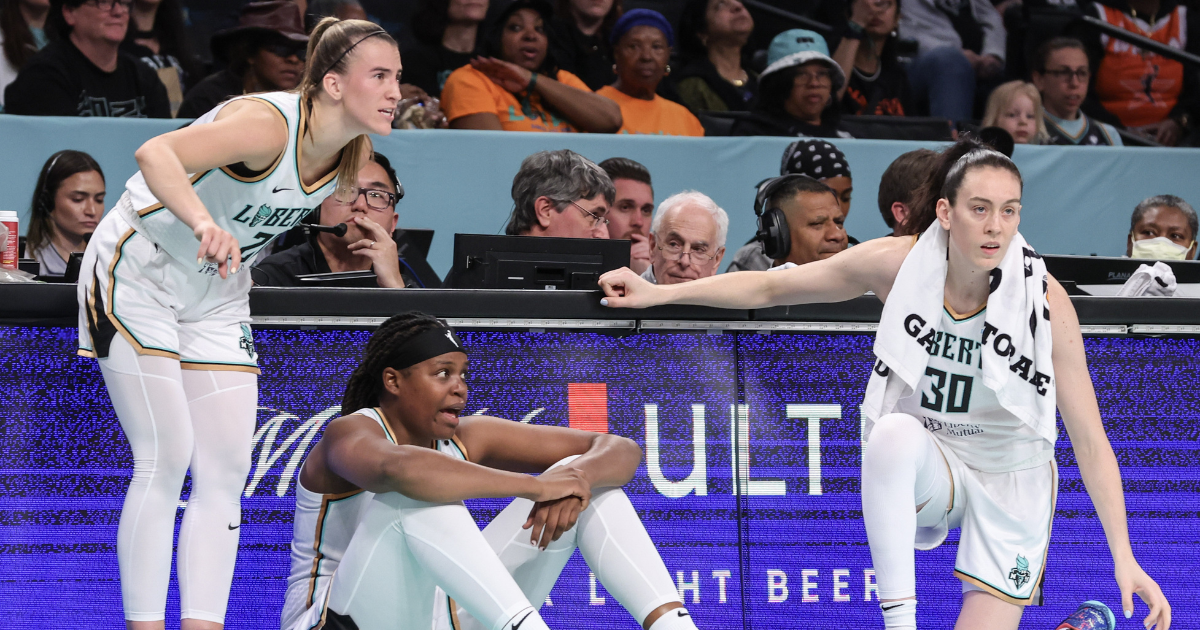 Sabrina Ionescu, Jonquel Jones and Breanna Stewart all wait at the scorers table to check into a game. Ionescu is standing, Stewart is kneeling, and Jones is seated between them with her elbows resting on her knees.