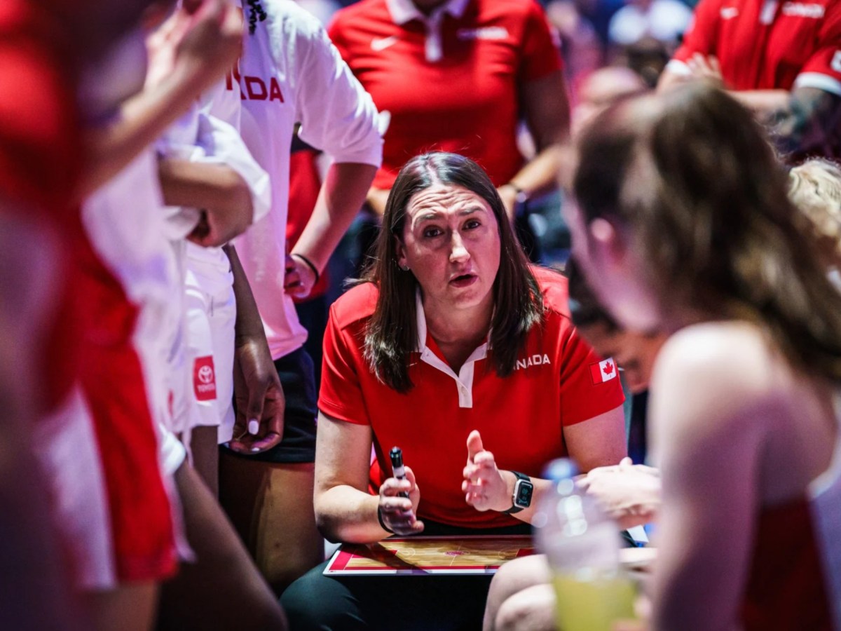 Carly Clarke speaks to Team Canada in a huddle. Players and coaches surround her as she talks.