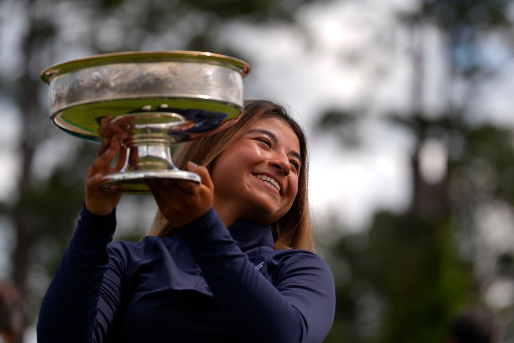 Maria Jose Marin hoists the Augusta National Women's Amateur trophy to her right and tilts her head left. She smiles off to the right at a different camera.