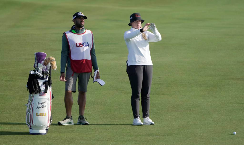 Ingrid Lindblad stands in the fairway and holds up her hands to visualize a landing spot for her golf shot. Her caddy and golf bag stand to the left of her.