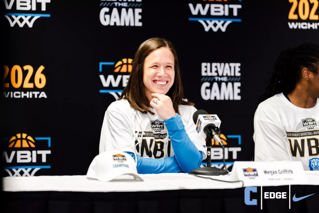 Columbia head coach Megan Griffith sits at a podium and rests her left hand under her chin. She is smiling wide and looking straight ahead. The WBIT logo is visible on the microphone in front of her and the backdrop behind her.