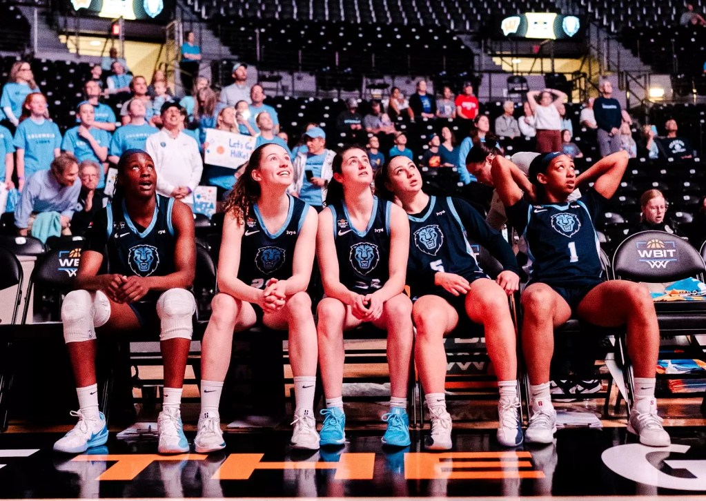 Five Columbia players sit on the bench and look up toward the video board, which is not visible in the photo. The players appear to be bathed in a reddish glow, and several Columbia fans wearing light blue are visible in the stands behind them.