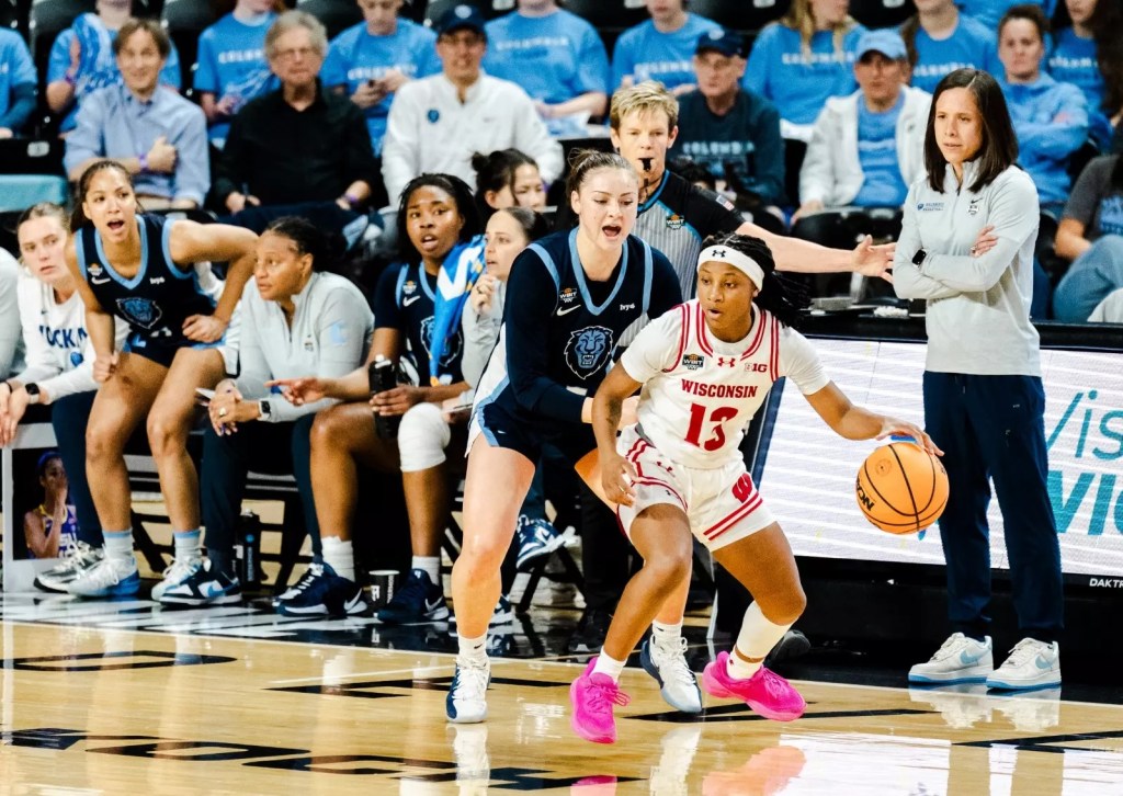Wisconsin guard Ronnie Porter dribbles with her left hand. She uses her body to shield the ball from Columbia guard Mia Broom, who is guarding her tightly right in front of the Columbia bench.
