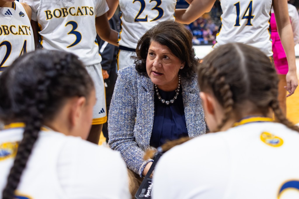 Quinnipiac head coach Tricia Fabbri sits during a timeout and looks at the players who are in the game. Several other players stand behind her with their arms around each other.