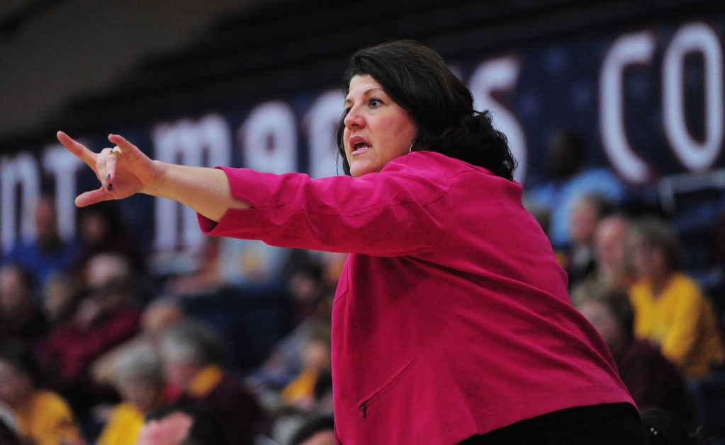 Quinnipiac head coach Tricia Fabbri is shown on the sideline wearing a bright pink blazer. Her left arm is outstretched, like she's pointing something out to the players on the court.