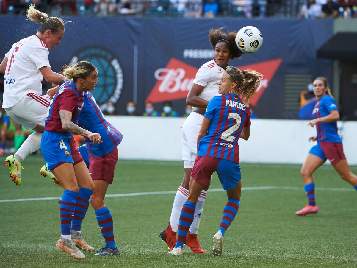A player in white is in the air after she has headed the ball over players in blue and red. Another player in white is behind the players in red and blue.