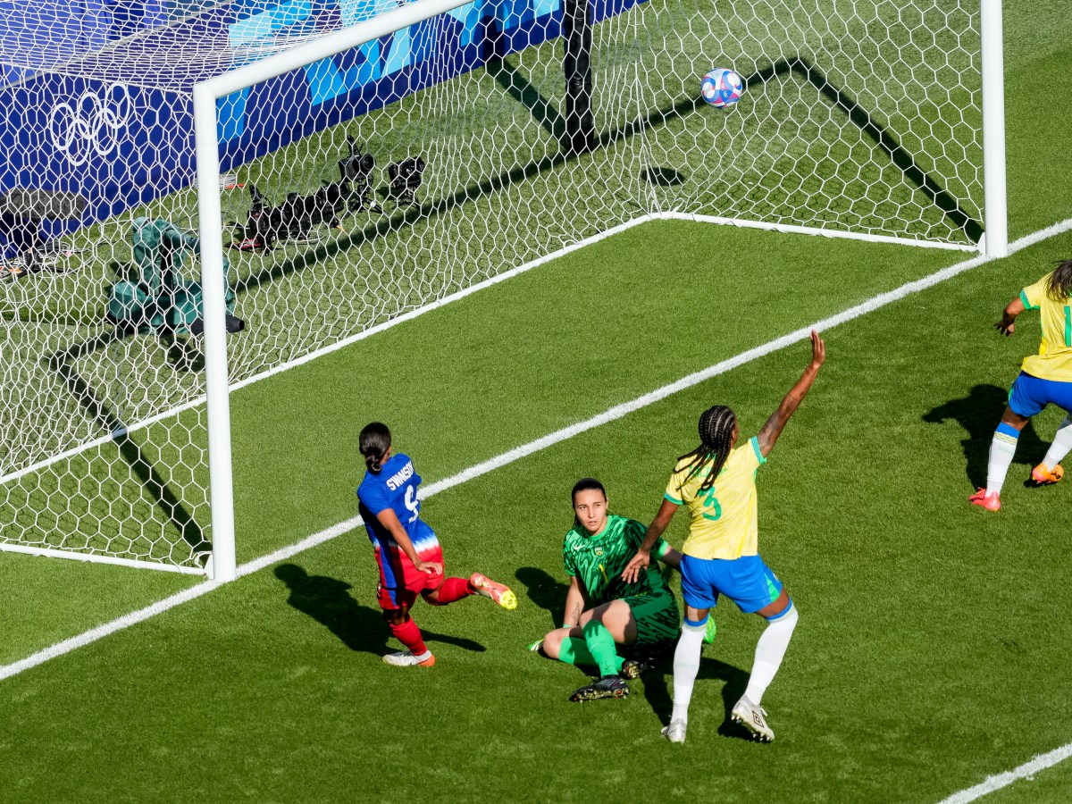 A player in blue watches a ball go into the net while two players in yellow raise their hands. A goalkeeper in green is on the ground.
