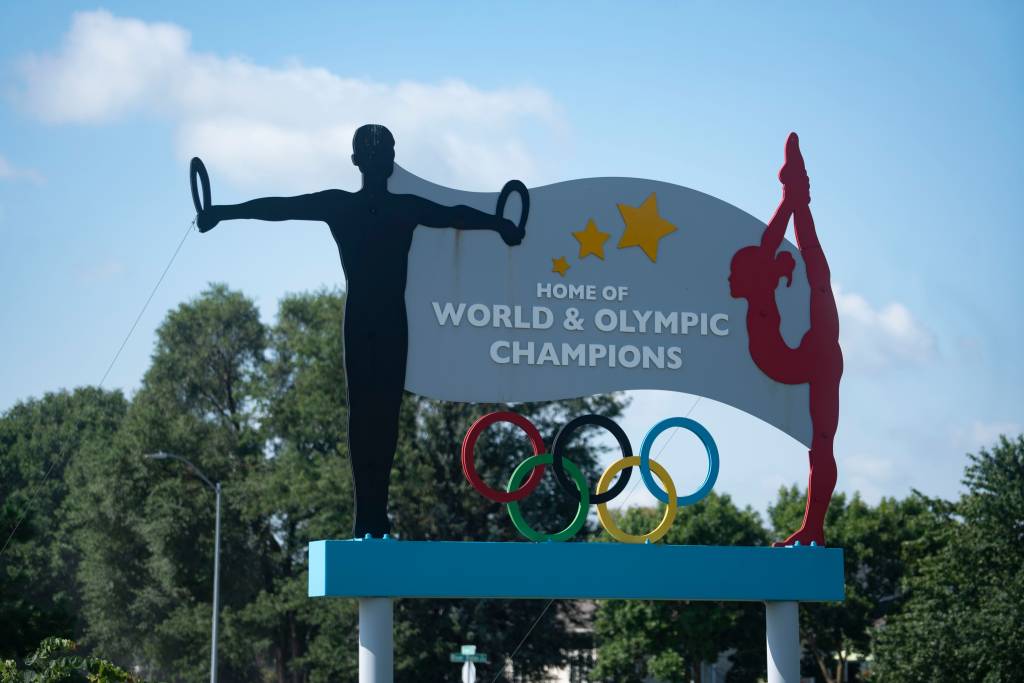 A sign featuring silhouettes of gymnasts and Olympic rings stands in front of a row of trees