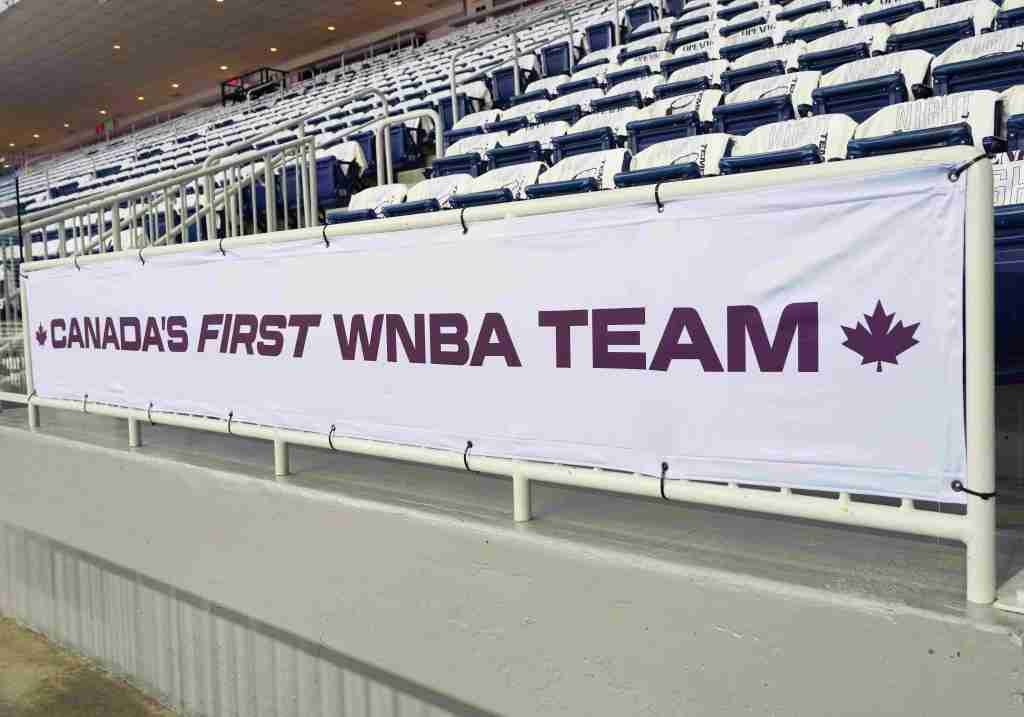 A banner that reads "Canada's first WNBA team" is zip-tied to a railing. Behind the railing, empty seats have white Toronto Tempo T-shirts draped over them.
